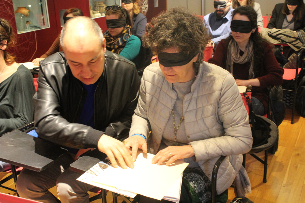 Due partecipanti al workshop leggono con le mani una riproduzione tattile. Sulla sinistra c’è una persona con disabilità visiva e sulla destra una bendata