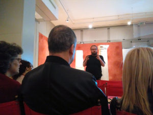 Gian Maria Greco in piedi spiega il funzionamento delle audio descrizioni. Davanti a lui, ripresi di spalle, alcuni dei partecipanti al workshop, seduti